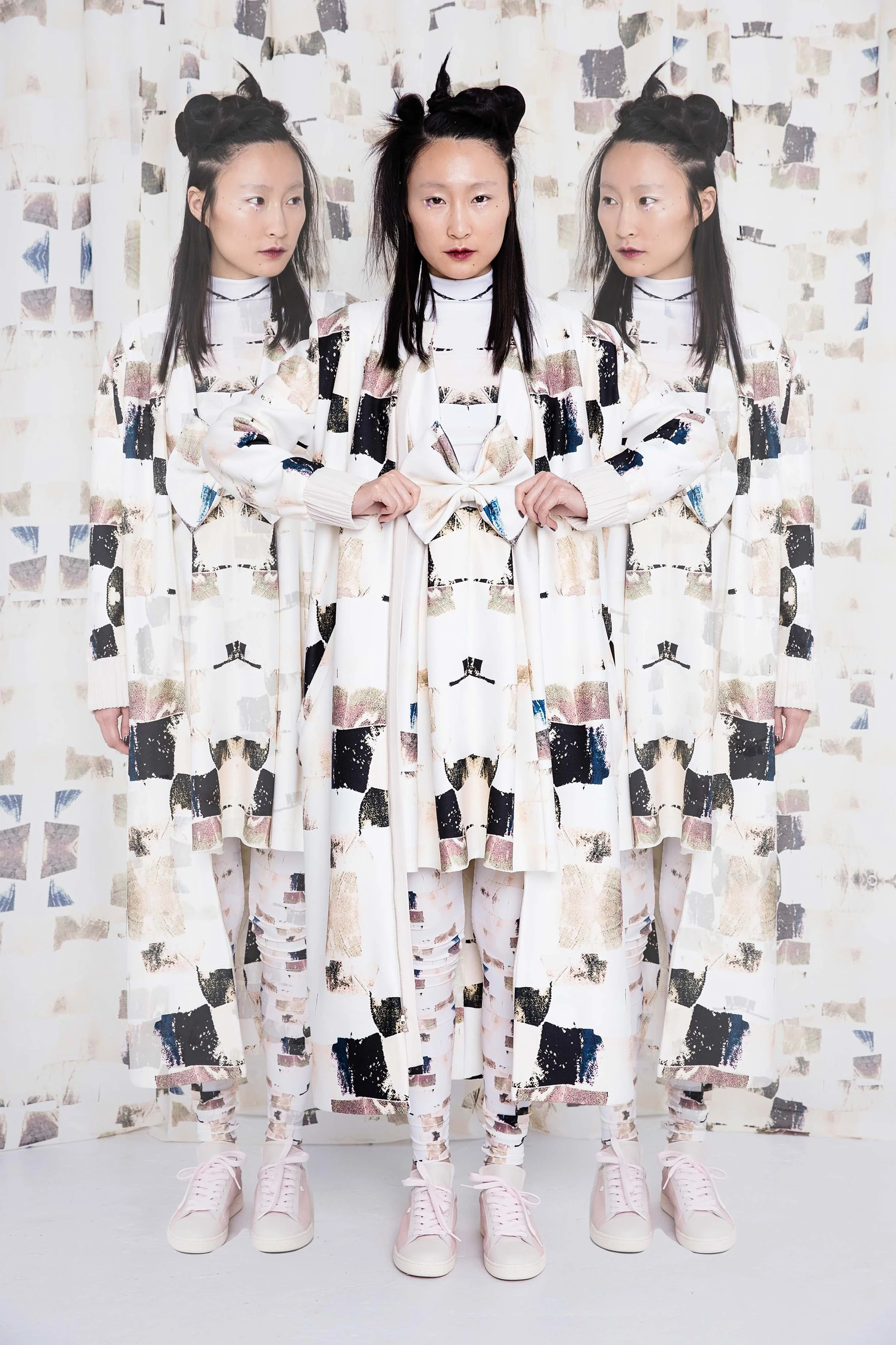 Three women with identical hairstyles and patterned clothing standing against a matching geometric background, with the central woman adjusting her outfit.