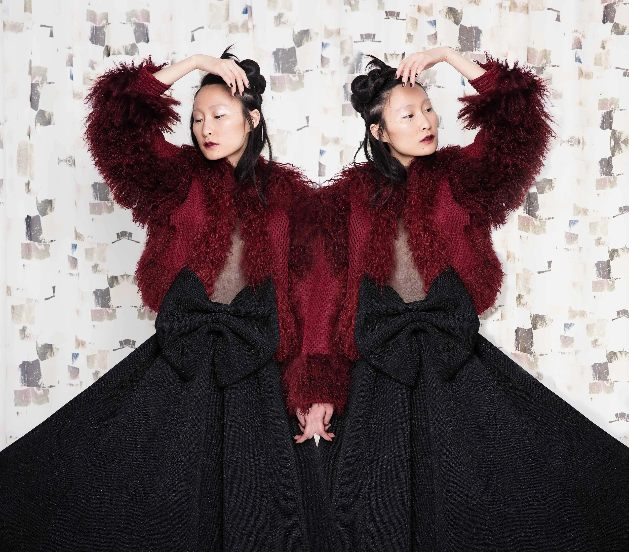 A mirrored image of a woman in a red textured sweater with large black bows on her black skirt, posing with one hand on her head and the other holding her skirt, standing in front of a white patterned background.
