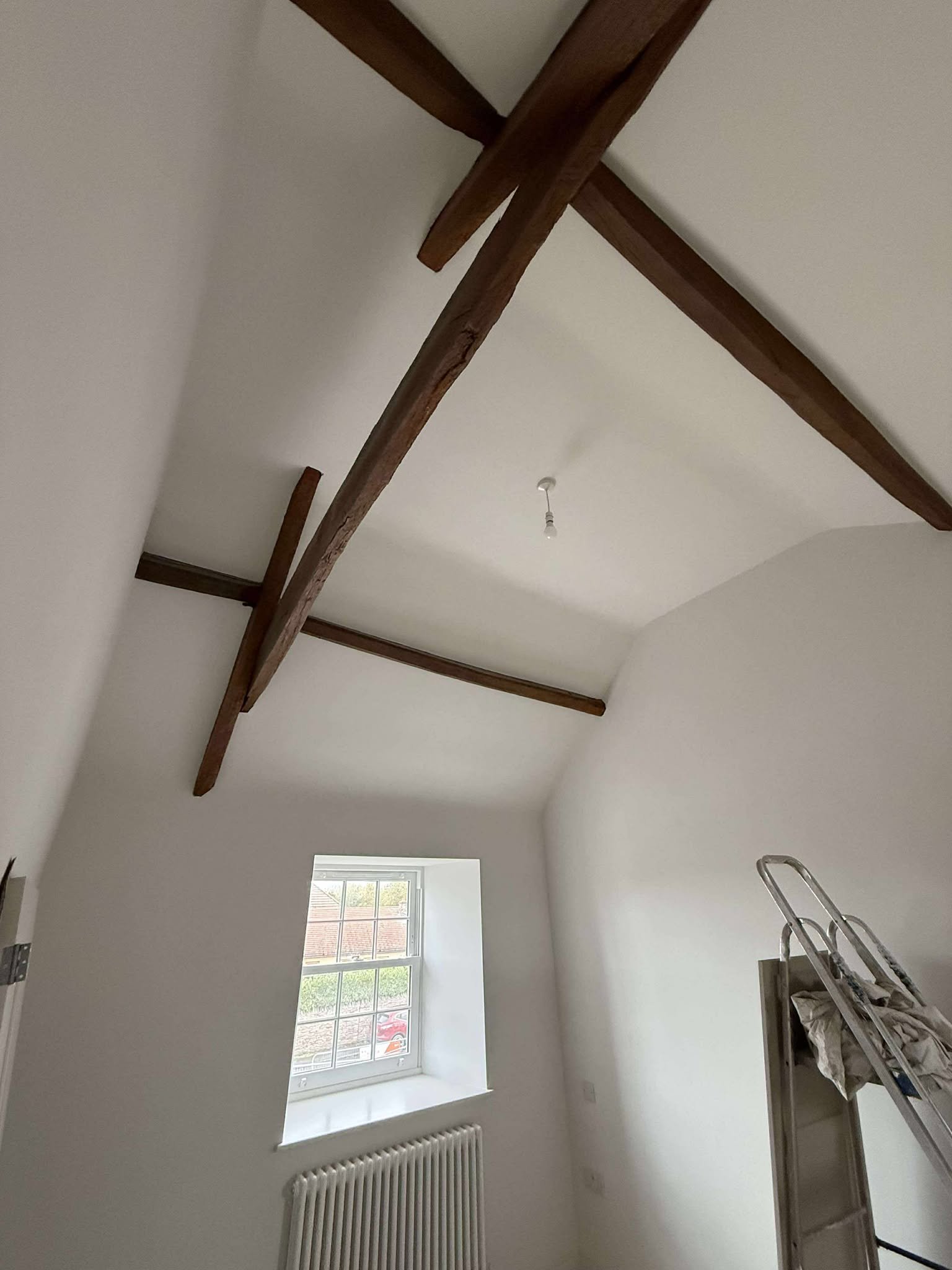 White room with exposed wooden ceiling beams, a small window, and a ladder.