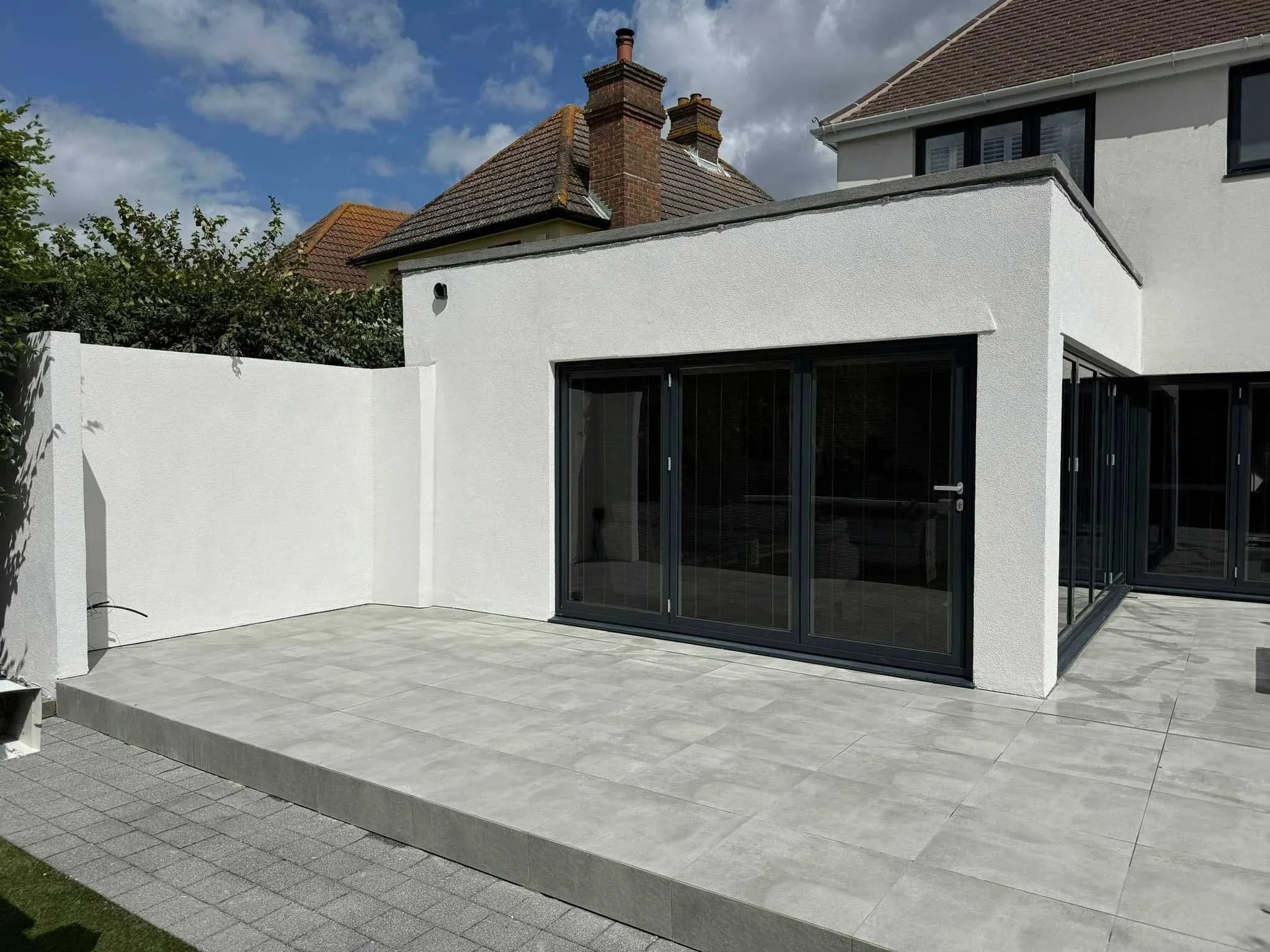 Modern house backyard with white stucco wall, large sliding glass doors, tiled patio, and neighboring houses with chimneys under partly cloudy sky.