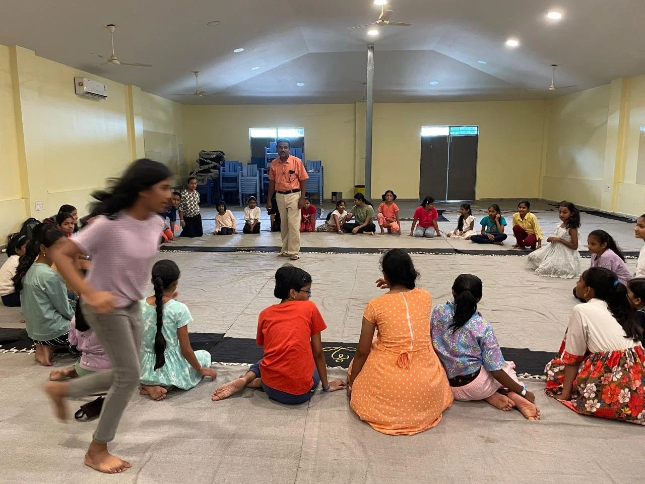 Children sitting in a circle on the floor with a man standing in the center in a large indoor hall, some children are watching while a girl walks in front of the group, all are casually dressed.