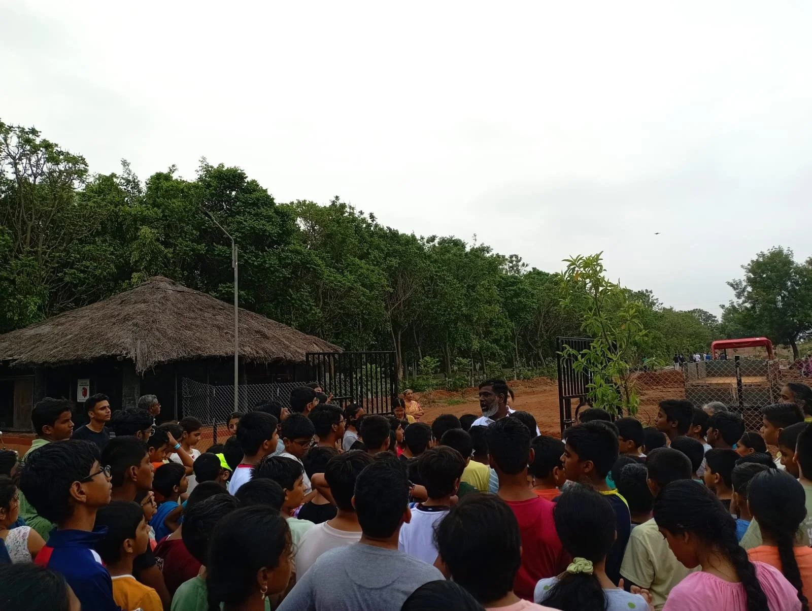 A large group of children and adults gathered outdoors in front of a gate, with greenery and trees in the background, possibly at a park or zoo.