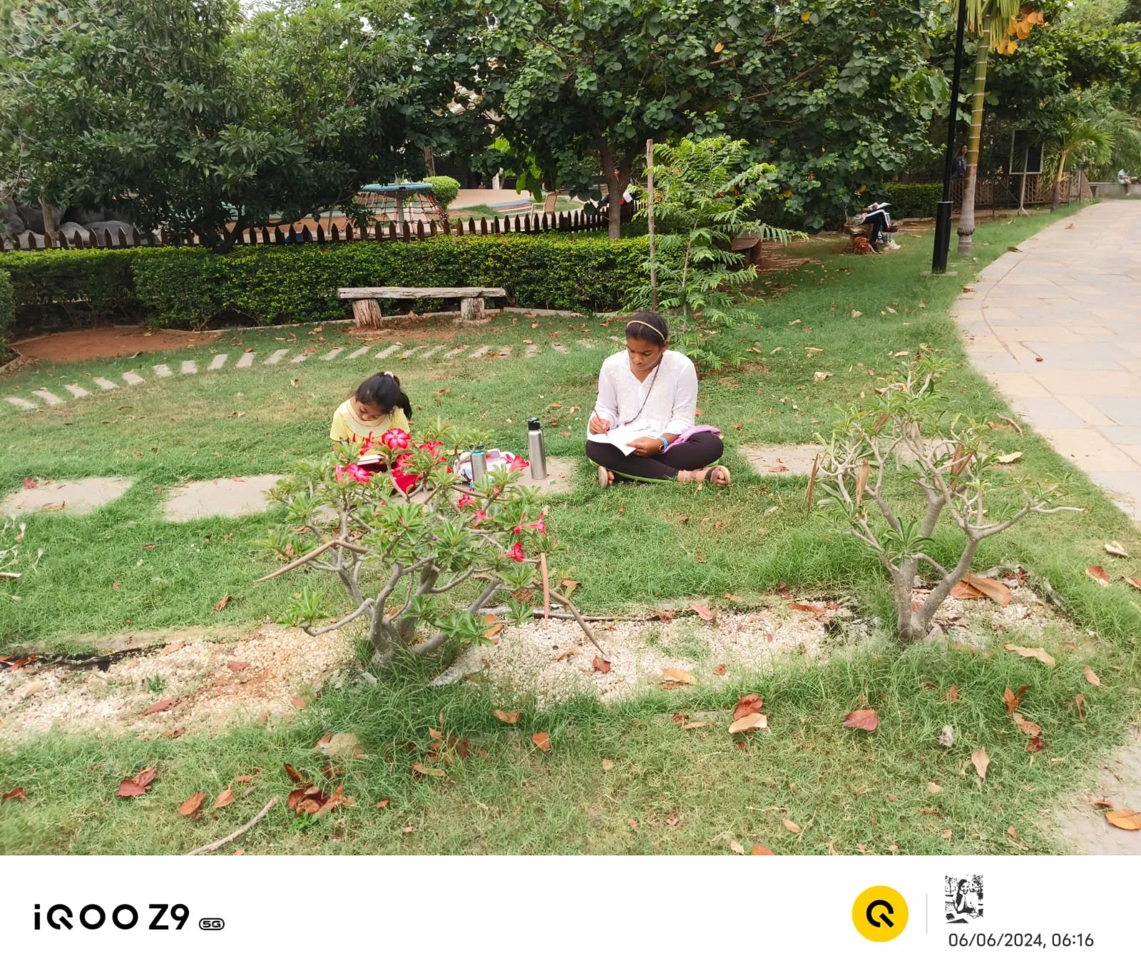 A woman and a young girl sitting on grass in a park, surrounded by plants and trees, with the woman writing in a notebook and the girl reading a book.