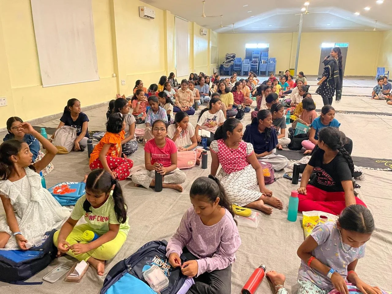 A large group of children sitting on the floor in a spacious room, listening attentively to a woman standing and speaking to them.