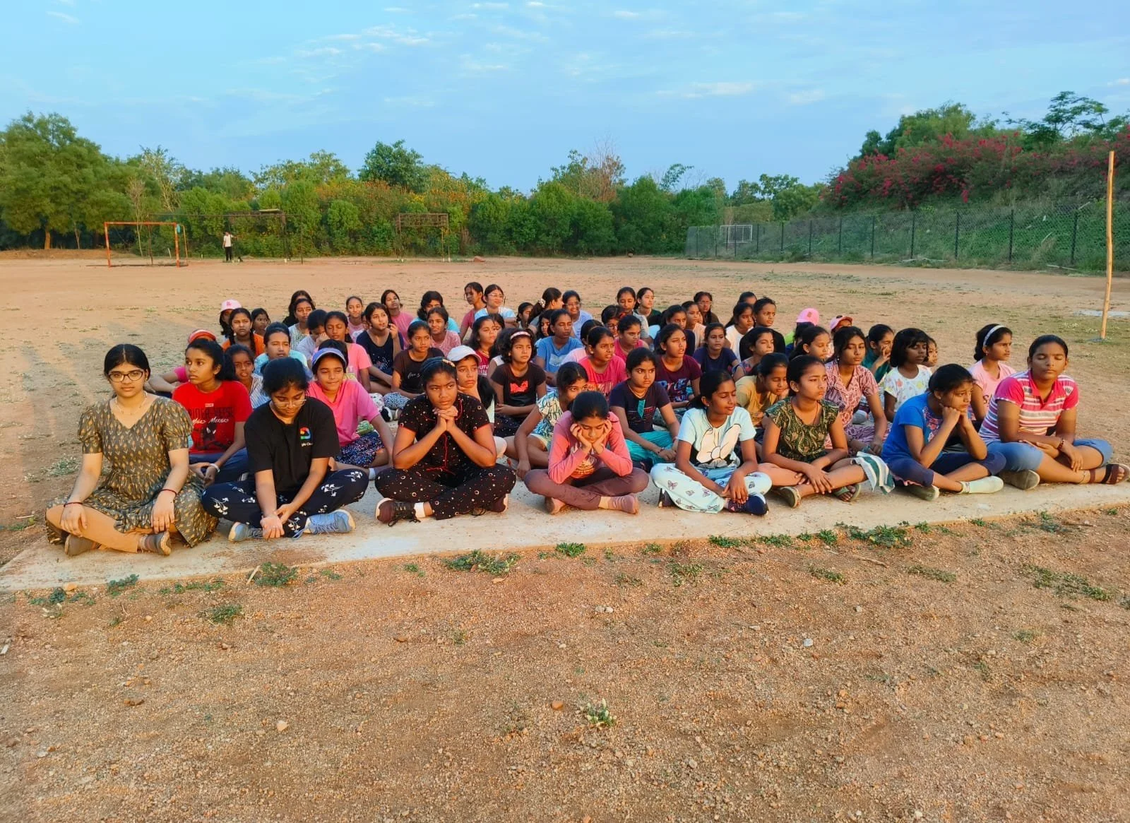 A large group of young girls sitting in a yoga pose on the ground outdoors, with a soccer field, trees, and a fence in the background.