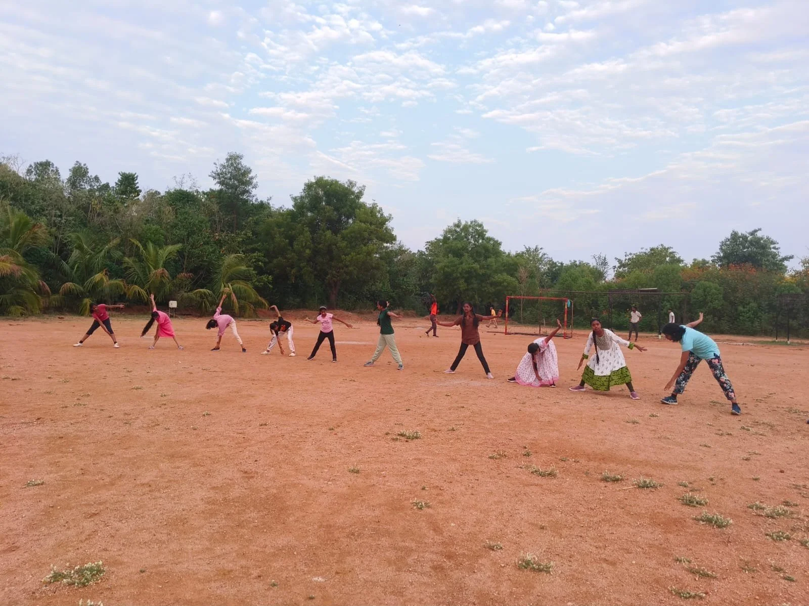 Group of children and young women playing on an open dirt field with trees and cloudy sky in the background.