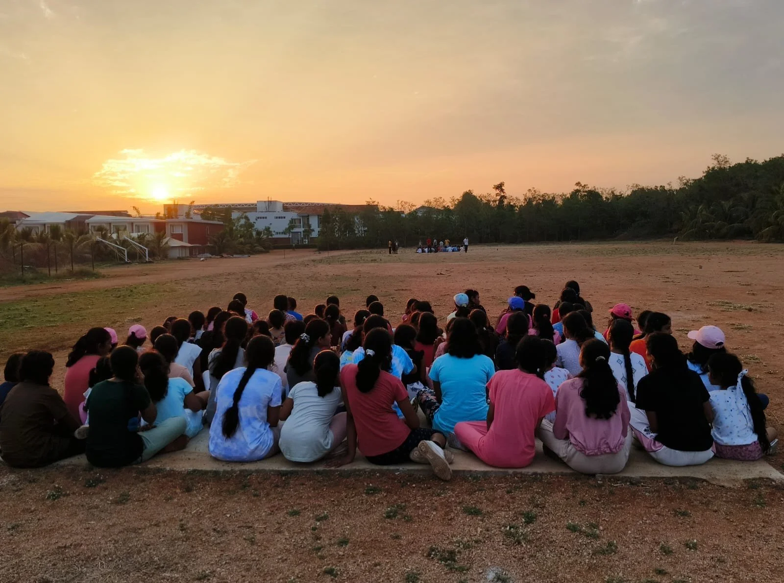 Group of children sitting on the ground facing a sunset, some wearing caps, in an open field with houses and trees in the background.
