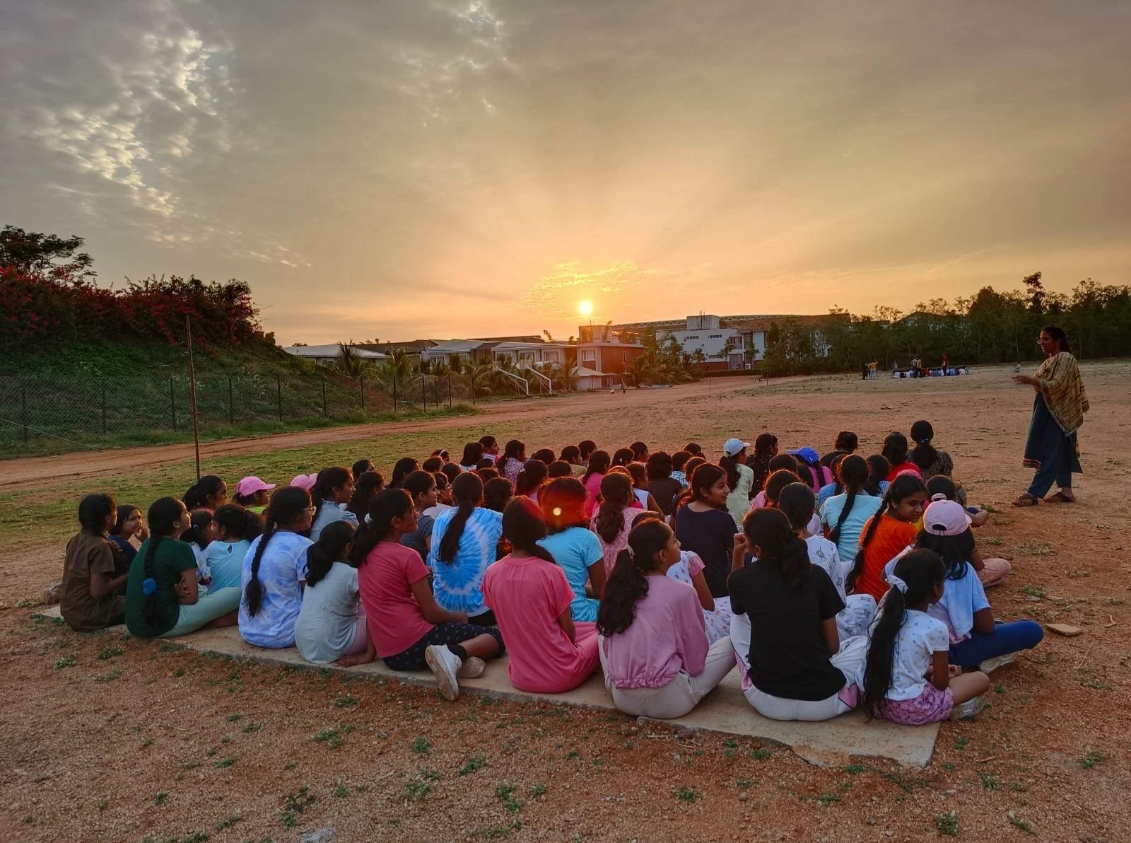 A group of children sitting on a mat outdoors during sunset, facing a woman standing in front of them on a dirt field.