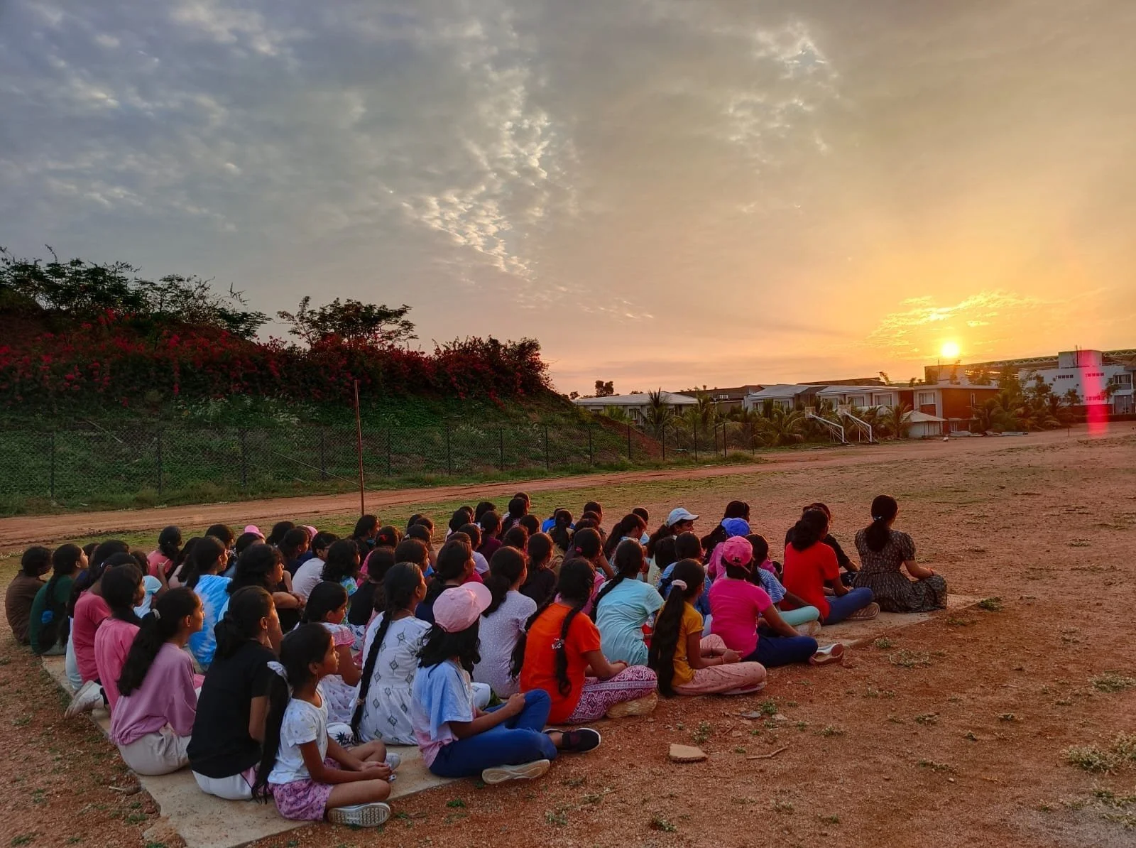 A group of children sitting on the ground outdoors at sunset, facing a field with trees and houses in the distance.