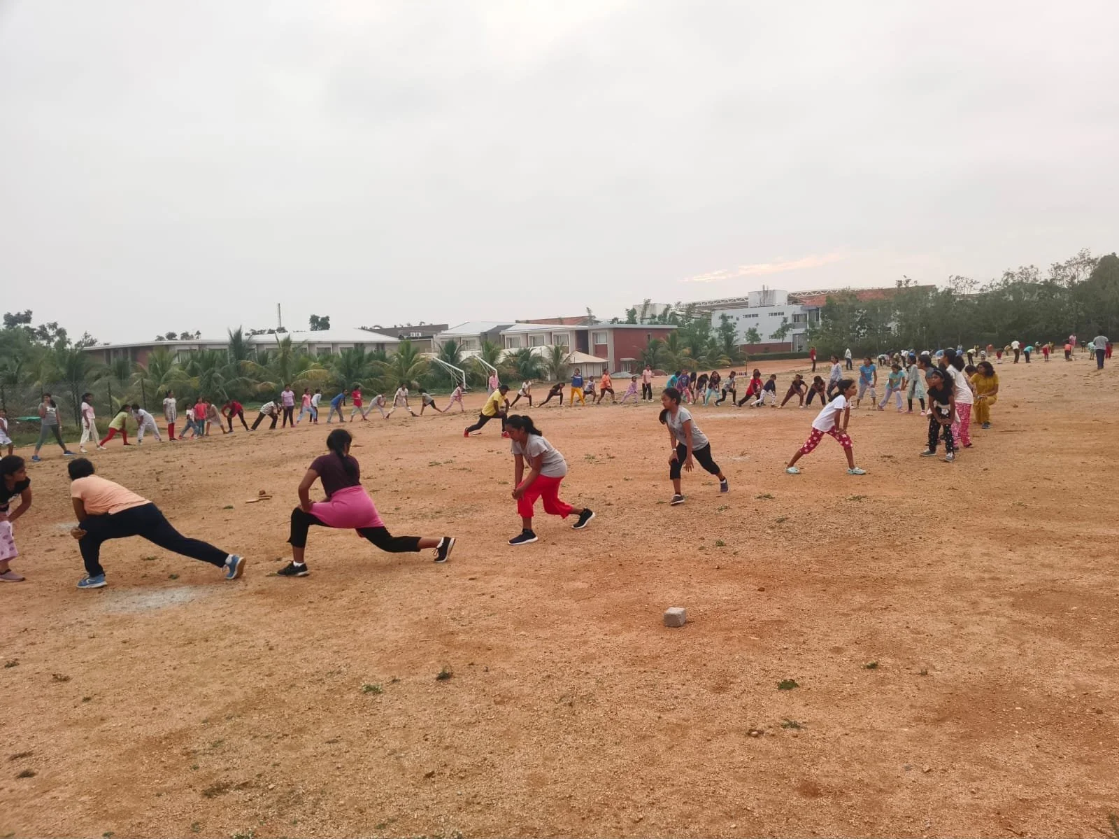 Children and adults playing tug-of-war and running in a large outdoor dirt field, with houses and trees in the background under cloudy skies.