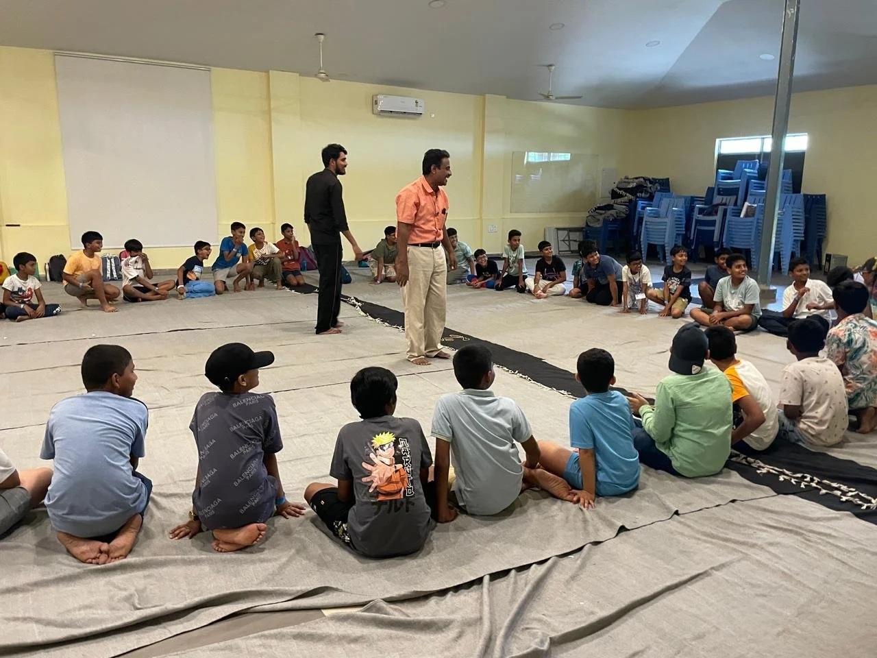 Children sitting in a circle on mats around two men standing in the middle of a large room, possibly for a group activity or discussion, with stacked blue chairs and a window in the background.