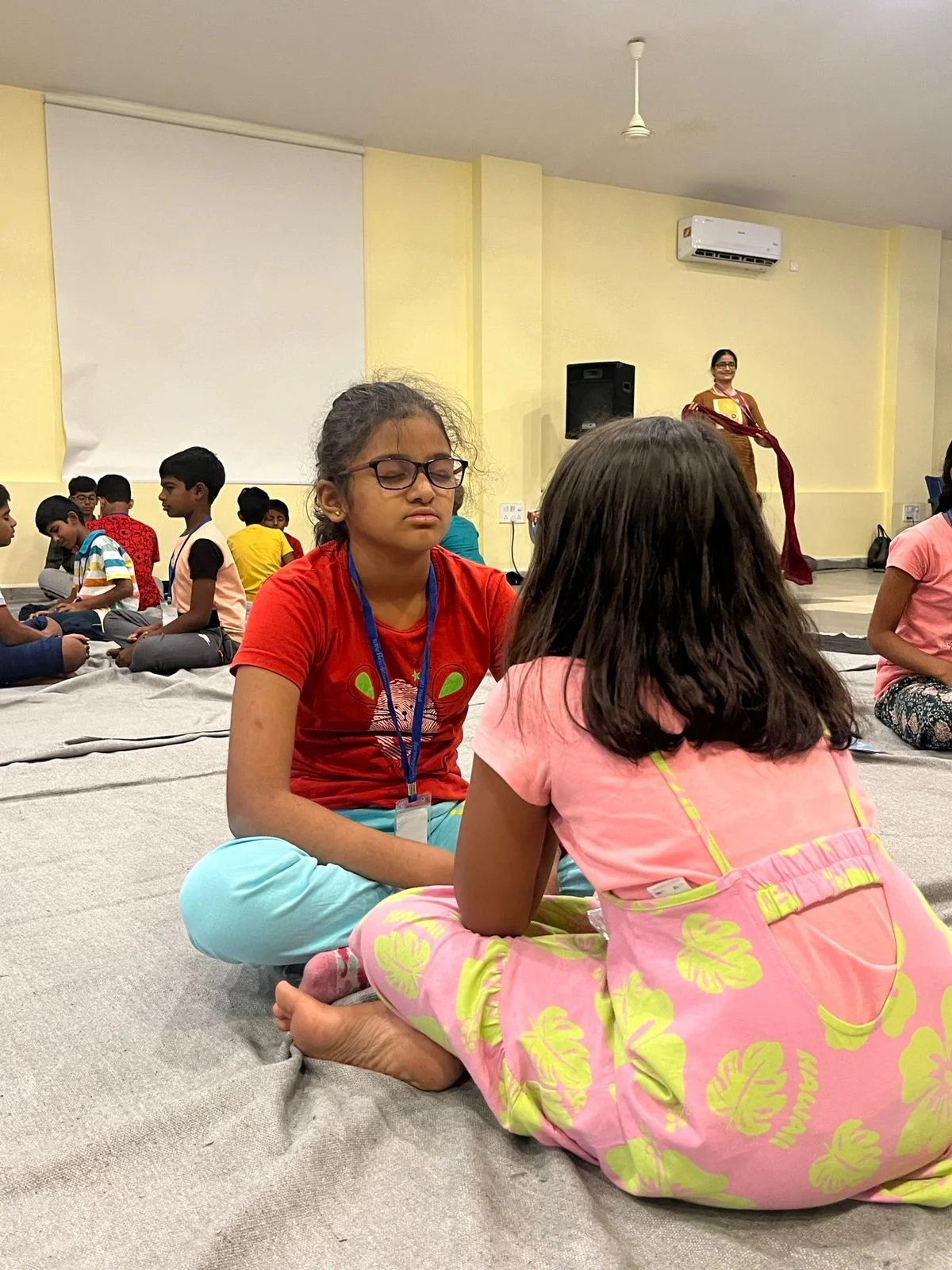 Children sitting cross-legged in a meditative pose with their eyes closed in a group setting indoors, with an adult woman standing in the background.