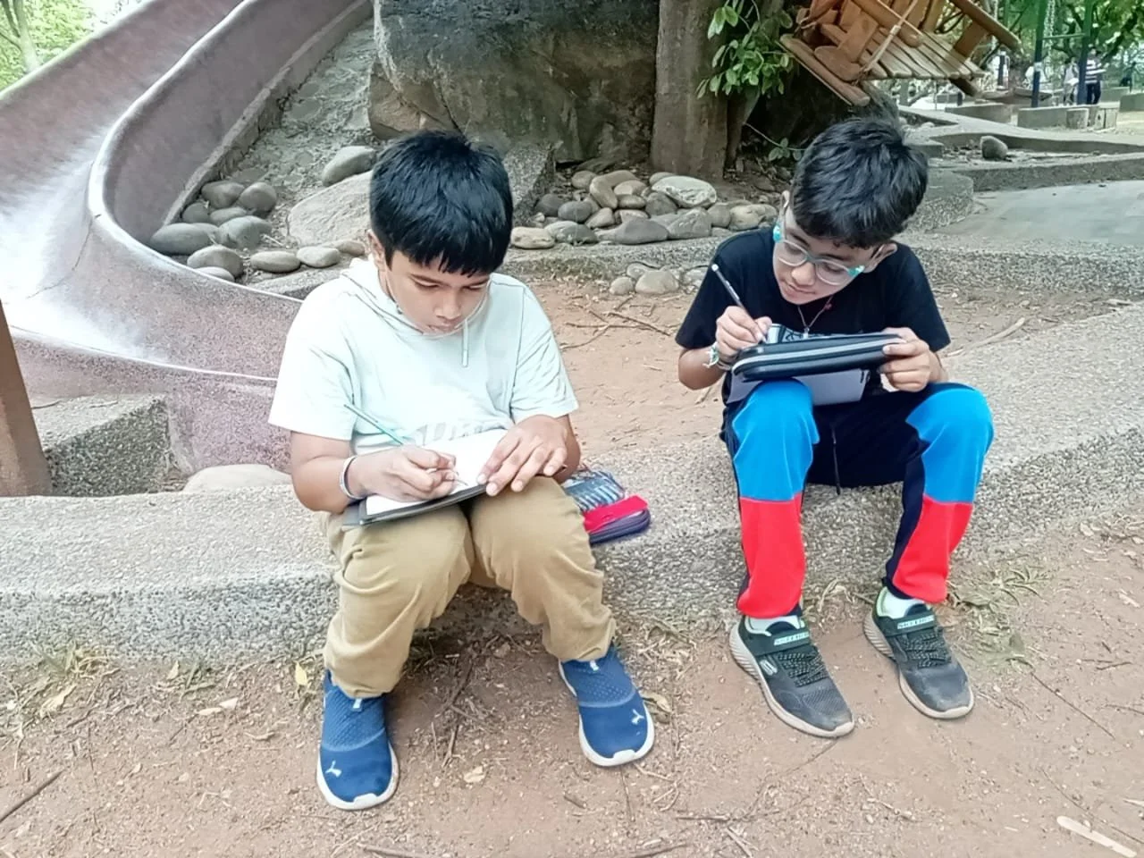 Two boys sitting on a concrete edge outdoors, each focused on drawing or writing in notebooks. One boy wears a white shirt and khaki pants, the other wears a black T-shirt and blue pants with red sections. There is a slide and small rocks behind them