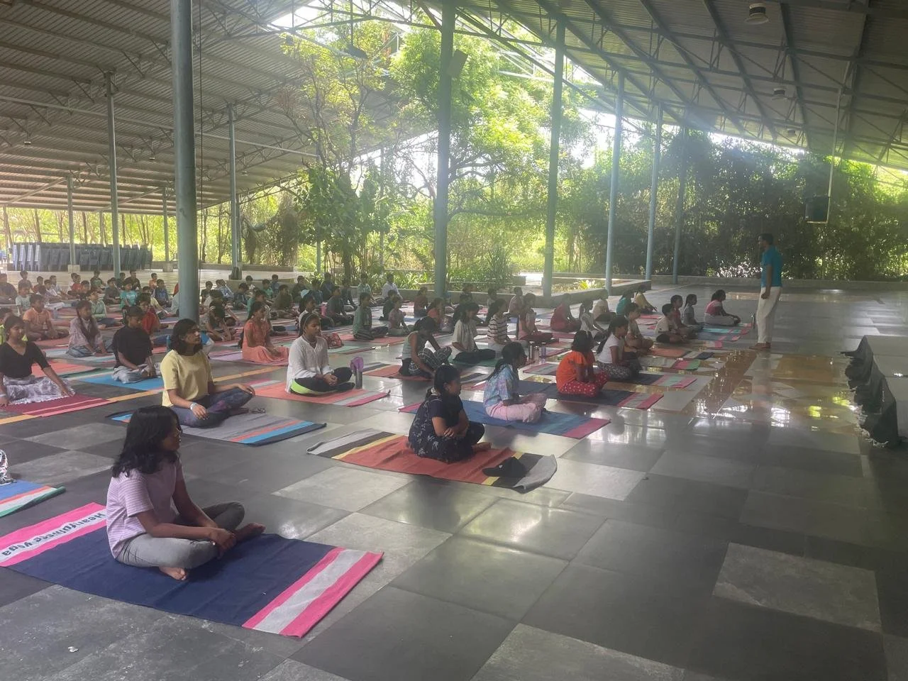 Group of women and children practicing yoga outdoors under a metal roof, sitting on mats in rows, with a teacher standing in front of them, some green trees in the background.