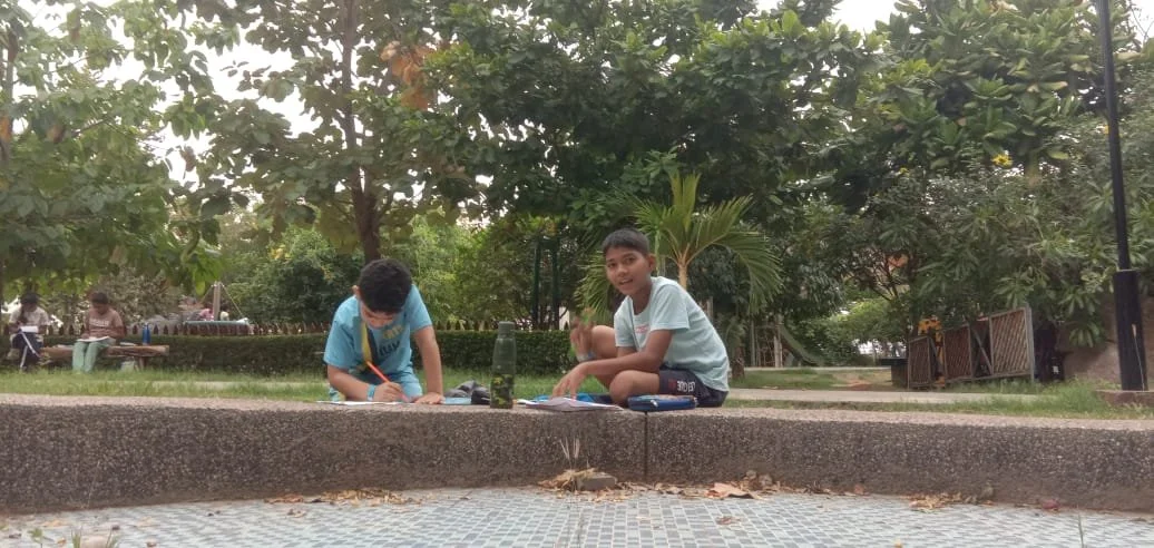 Two boys sitting on a park sidewalk, drawing on paper with trees and other people visible in the background.