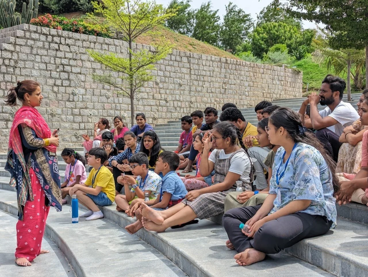 A woman in traditional attire standing and speaking to an outdoor audience of children and adults seated on stone steps during daytime with trees and a brick wall in the background.