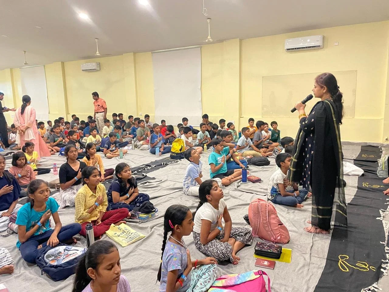 A woman in traditional black saree speaking into a microphone addressing children sitting on mats inside a classroom or hall.
