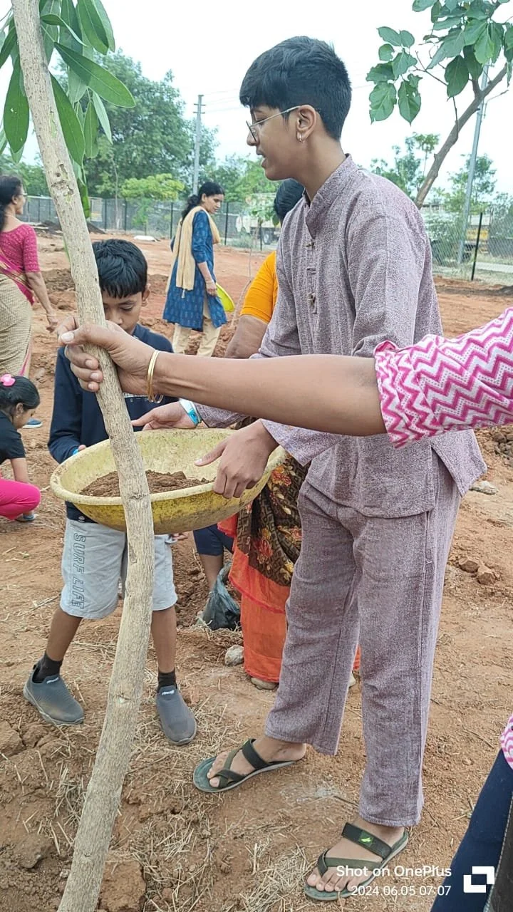 People planting a tree outdoors, with some children and adults involved in the activity.