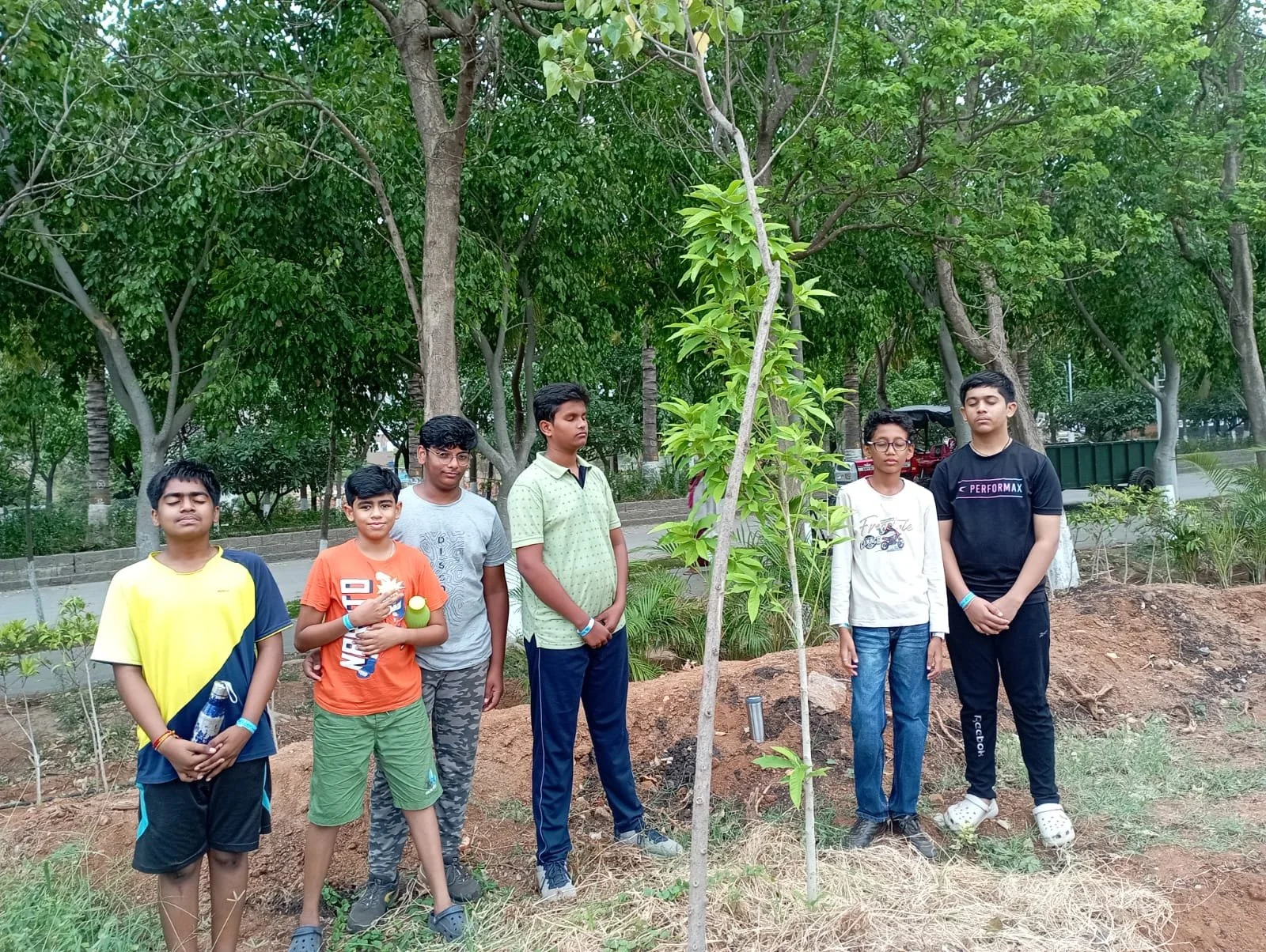 Six boys standing in a park near a young tree, with lush green trees in the background. Some boys are holding water bottles and the boys are standing on the soil.
