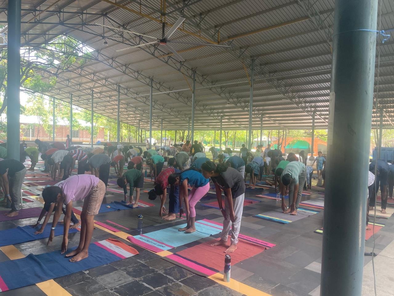 Group of people participating in a yoga class under a covered outdoor space, practicing yoga poses on mats.