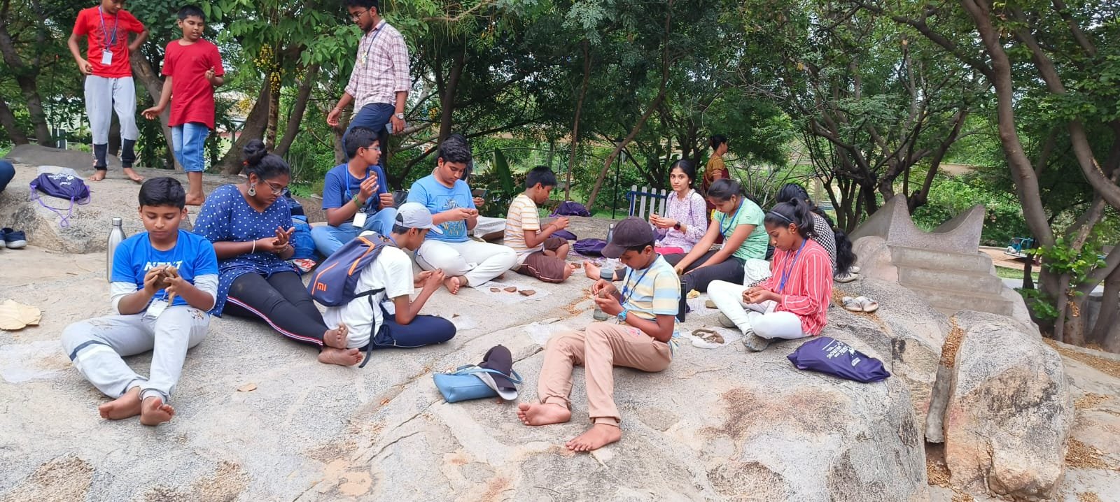 Group of children and adults sitting and relaxing outdoors on large rocks surrounded by green trees.