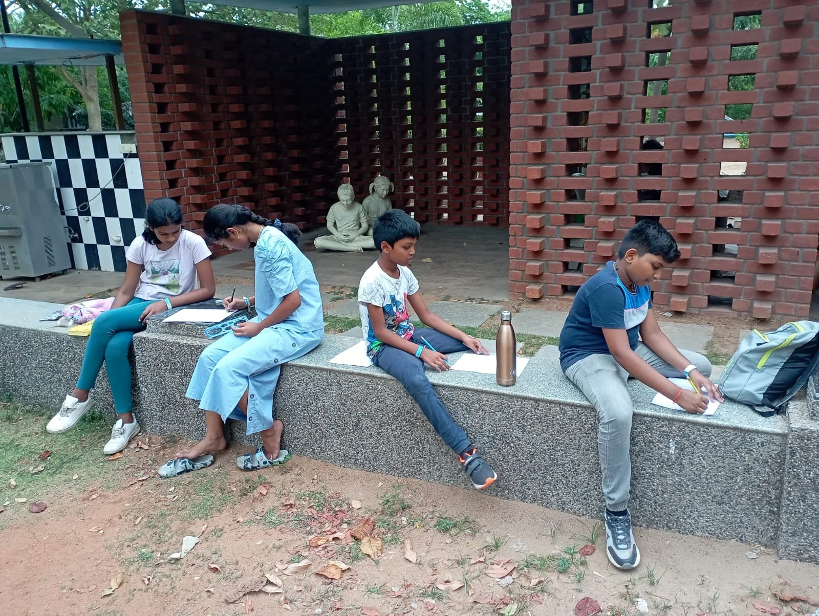 Four children sitting on a concrete bench outdoors, engaged in drawing or writing in notebooks, with trees and statues in the background.
