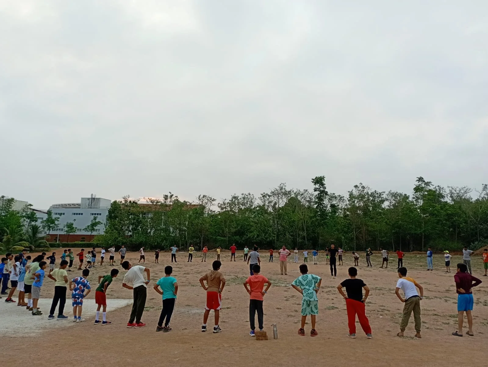 Group of children and adults standing in a large open field, forming lines for an outdoor activity, with trees and buildings in the background under a cloudy sky.