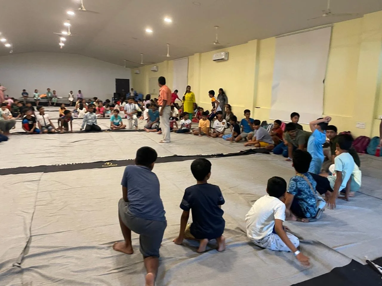 Children sitting on a large indoor floor, watching a performance or activity in a spacious hall with light yellow walls and ceiling fans.