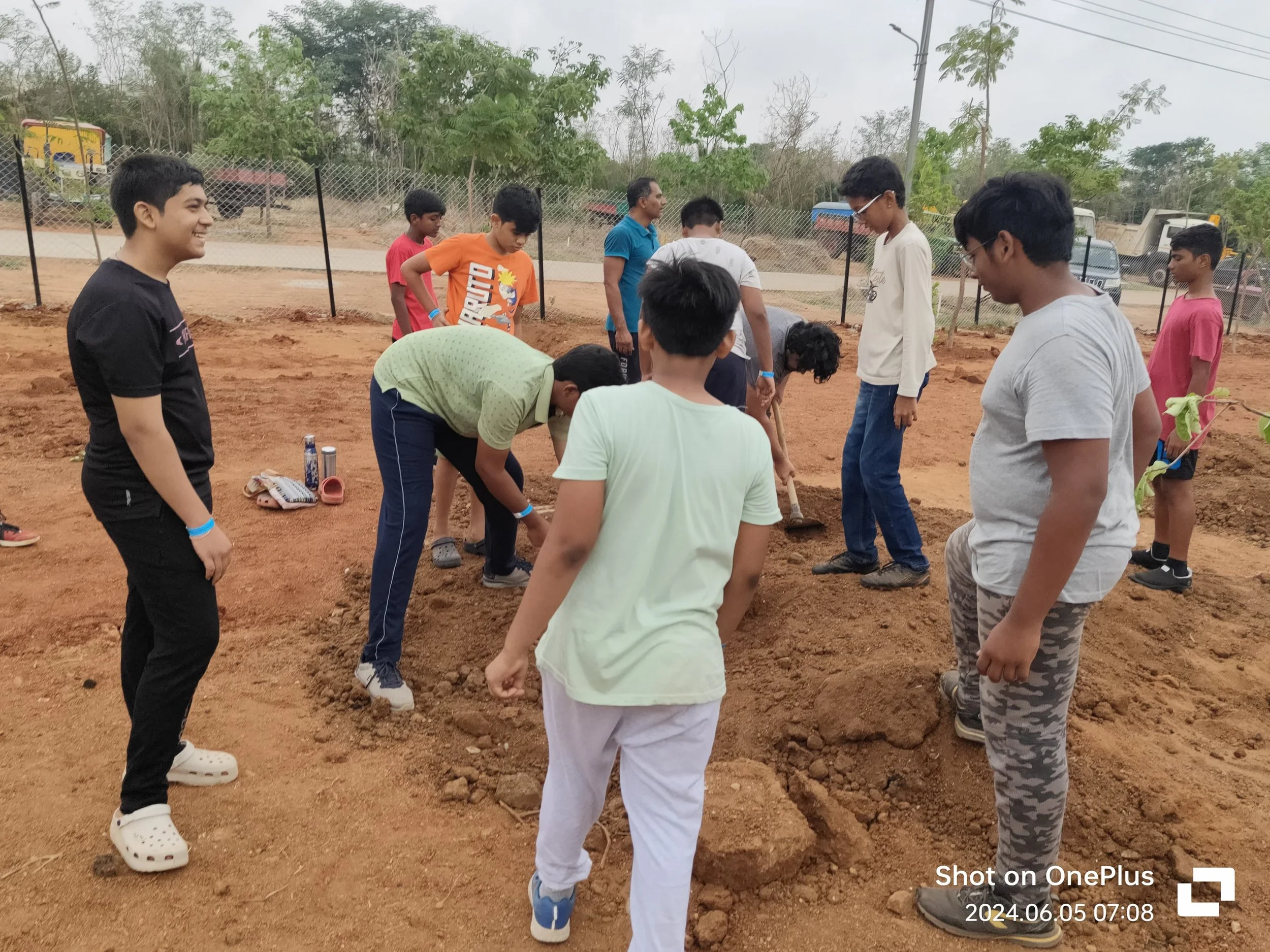 Group of children and teenagers planting a tree in an outdoor area with dirt and a wire fence in the background.