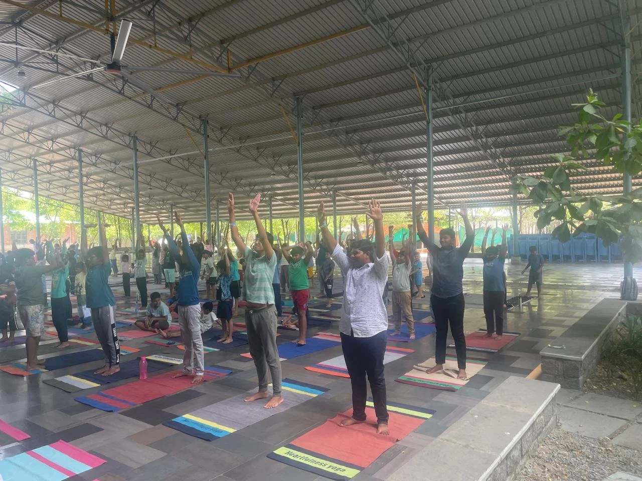 A large group of people participating in an outdoor yoga session under a shelter, standing on yoga mats with arms raised.