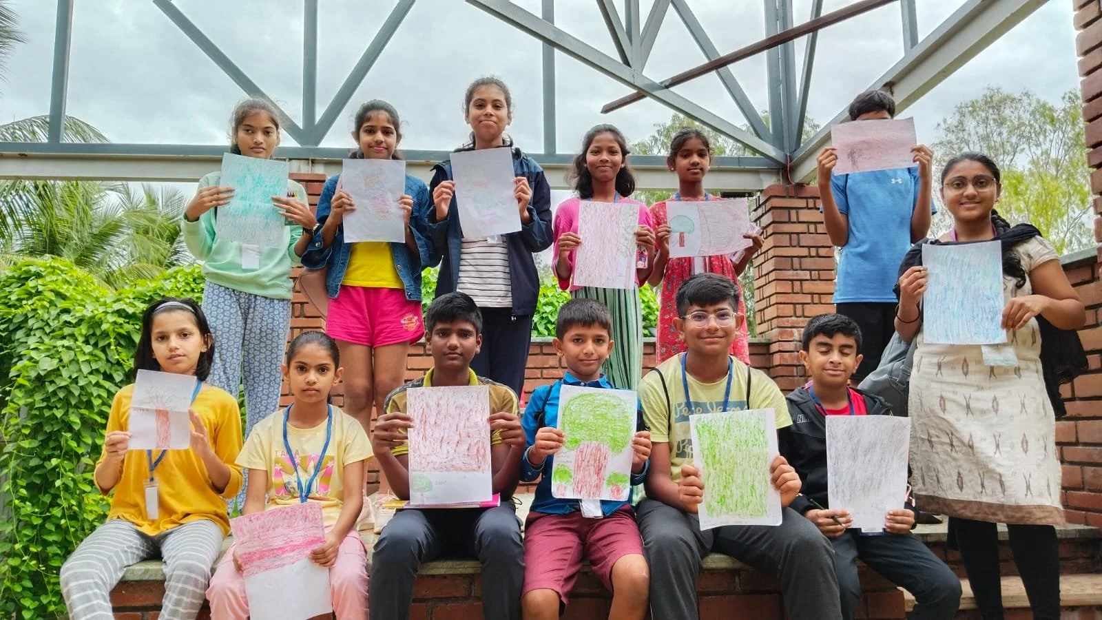 Group of children and teenagers posing outdoors, holding up their drawings of trees, in a setting with green foliage and a brick structure.