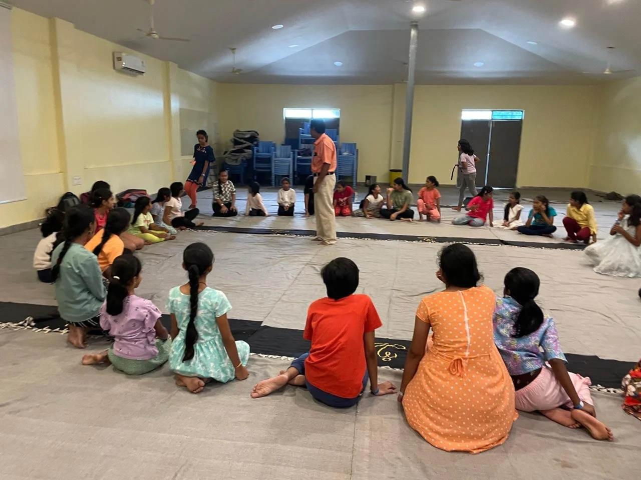 Children sitting cross-legged on the floor in a circle in a large indoor space, with adults standing and walking around, possibly during a cultural or educational event.