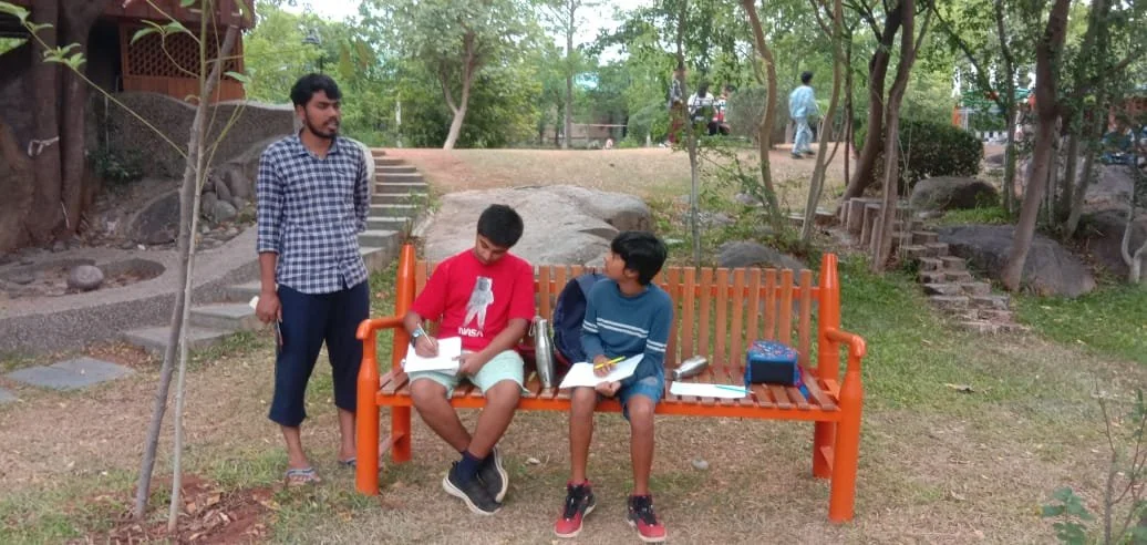 Two boys sitting on an orange bench with notebooks, and a man standing nearby in a park with trees and some steps in the background.