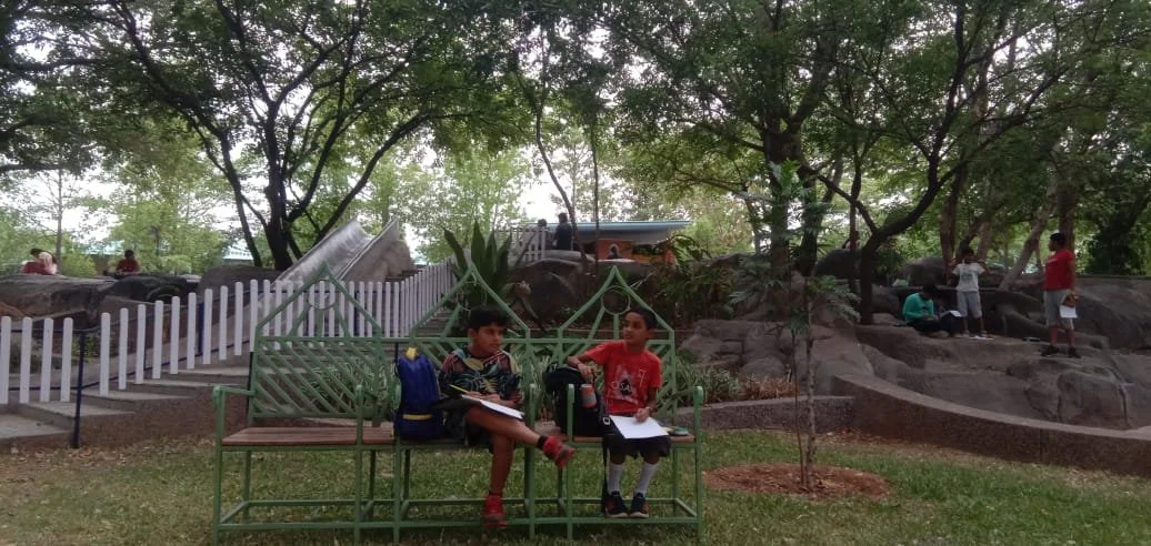 Two boys sitting on a green metal bench in a park, talking and with notebooks. Behind them are trees, rocks, and other children exploring. There is a small white fence and a see-saw in the background.