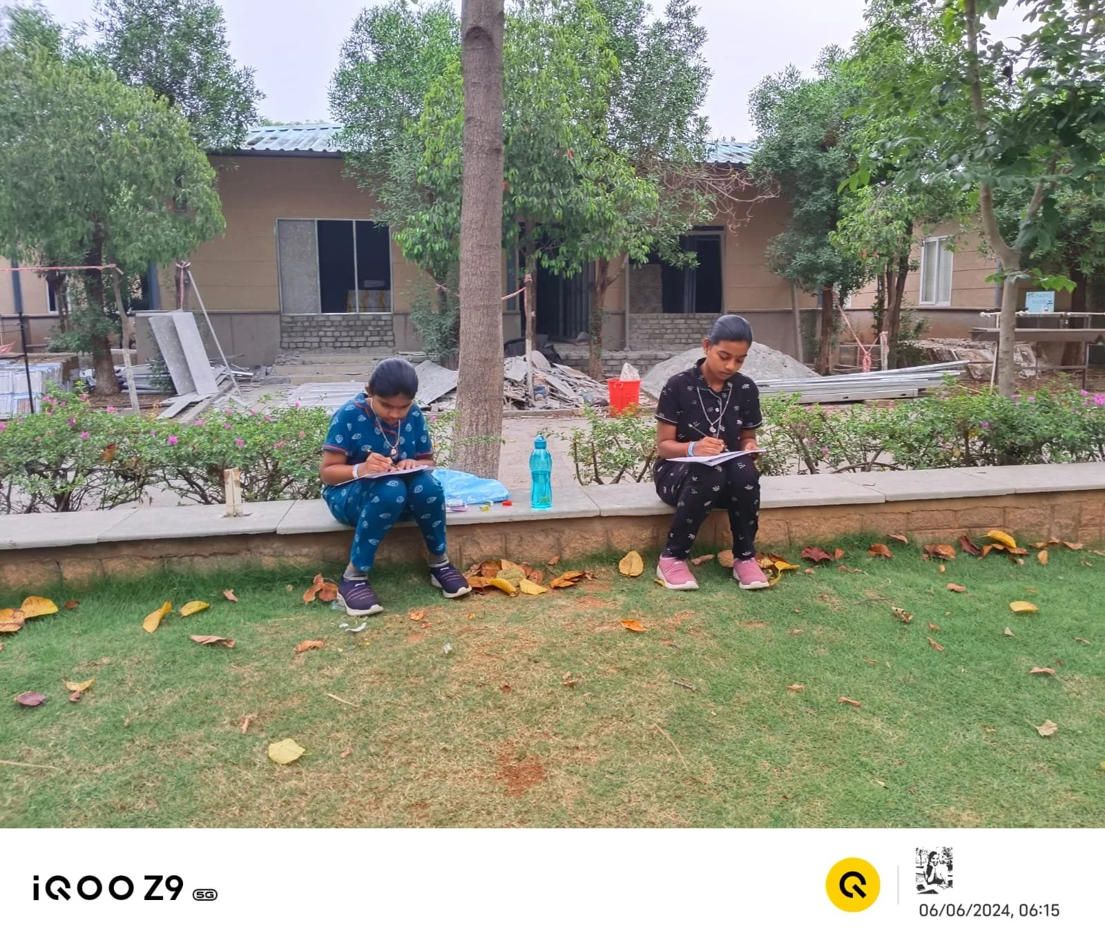 Two children sitting on the sidewalk, writing or drawing on notepads, with a background of trees and a building under construction, leaves scattered on the grass in front.