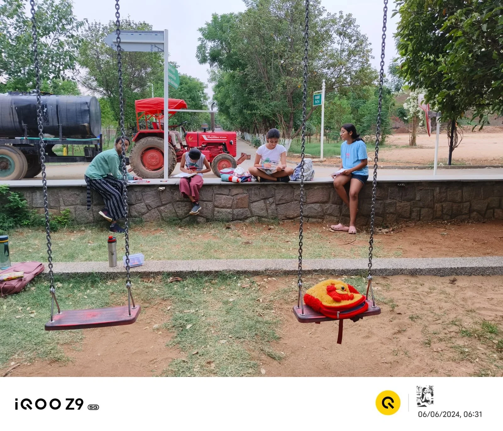 Four children sitting on a stone ledge in a park, two girls reading notebooks, and a boy looking at a phone. A red tractor and trees in the background, with a swing set in the foreground.