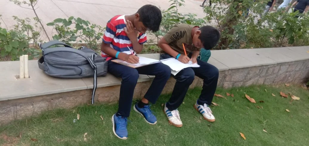 Two boys are sitting on a stone bench in a park, writing in notebooks. A backpack is beside them on the bench.