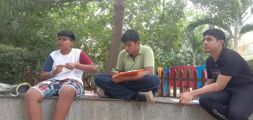 Three boys sitting outdoors on a concrete ledge, surrounded by trees and greenery, engaged in writing or drawing activities.