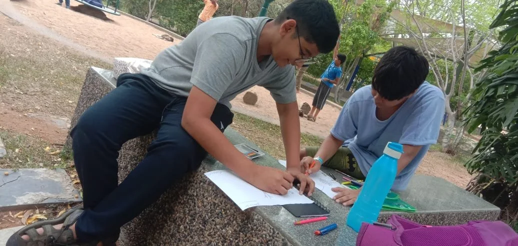 Two young men are sitting on a stone bench outdoors, working together on a large sheet of paper with pens and a smartphone, surrounded by trees and other people in the background.