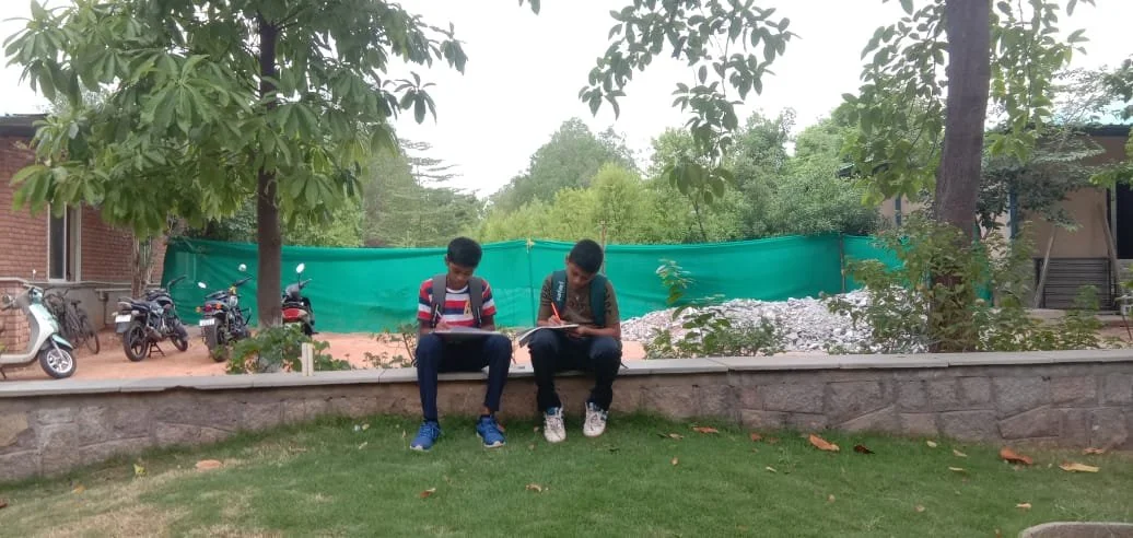 Two boys sitting on a stone wall under a tree in a park, with motorcycles and greenery in the background.