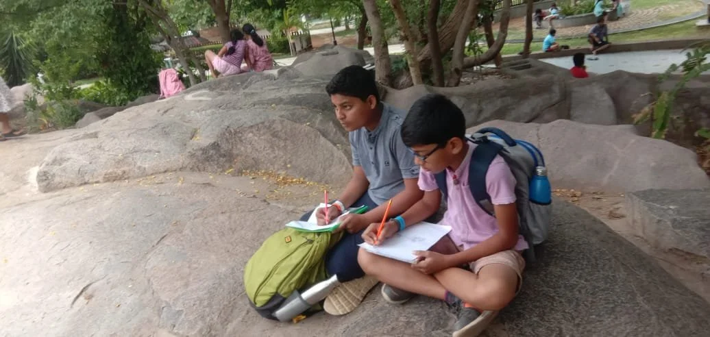 Two boys sitting on a large rock in a park, studying with notebooks and pens, with backpacks. Other children are playing and sitting in the background surrounded by trees.