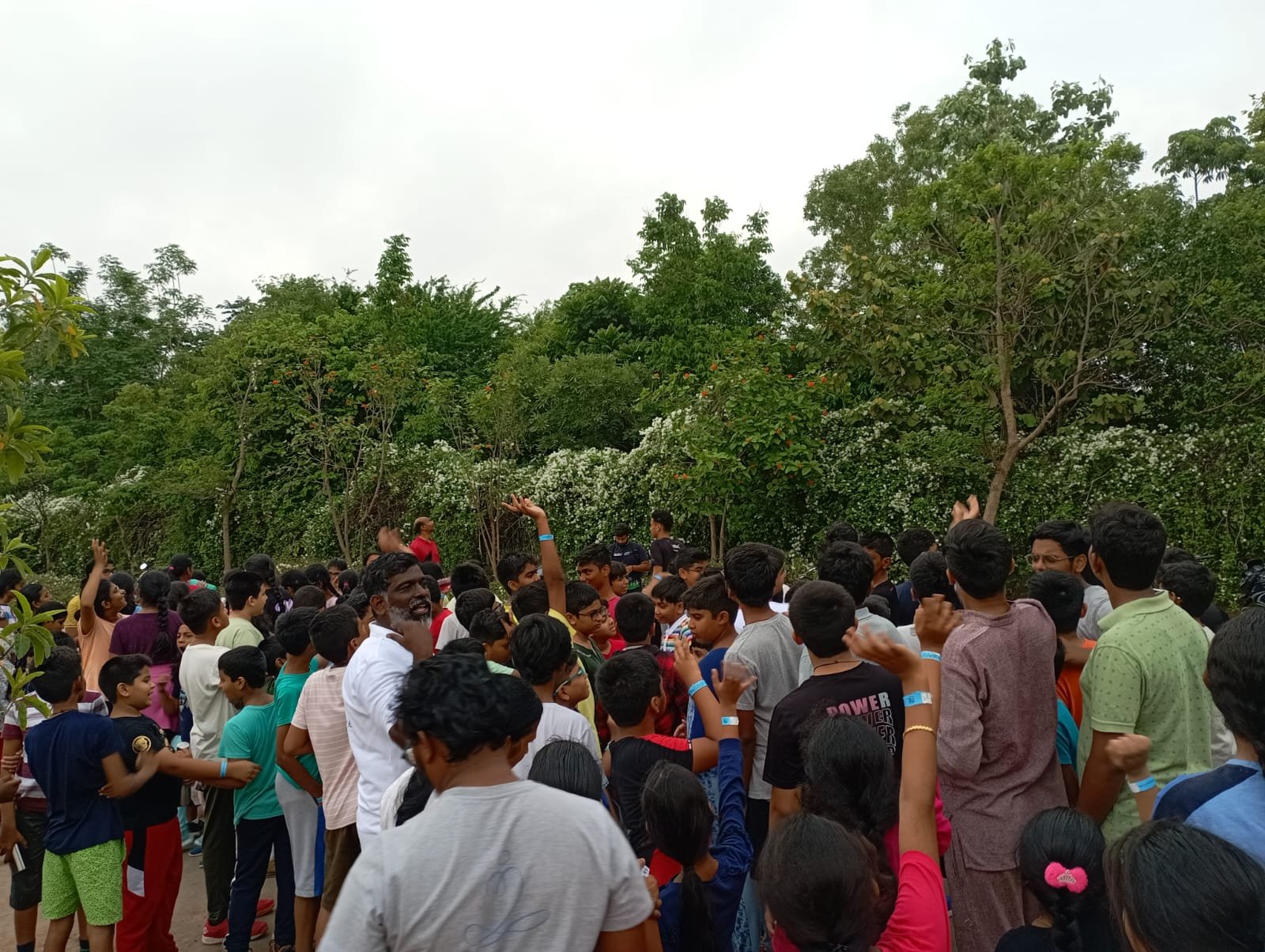 A large group of children and a few adults gathered outdoors among green trees, participating in an event or celebration.