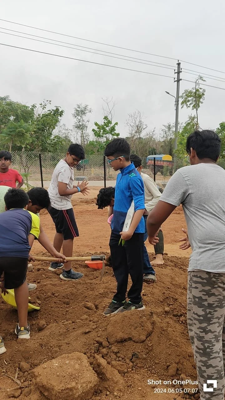 A group of children planting a small tree in a dug-up patch of soil outdoors, with a few adults helping. The children are wearing casual clothes and seem to be engaged in the activity.