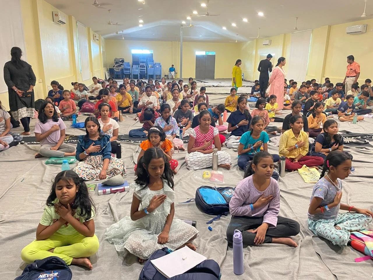 Children seated on the floor in a large hall, practicing meditation with eyes closed and hands on chest, guided by adults standing nearby.