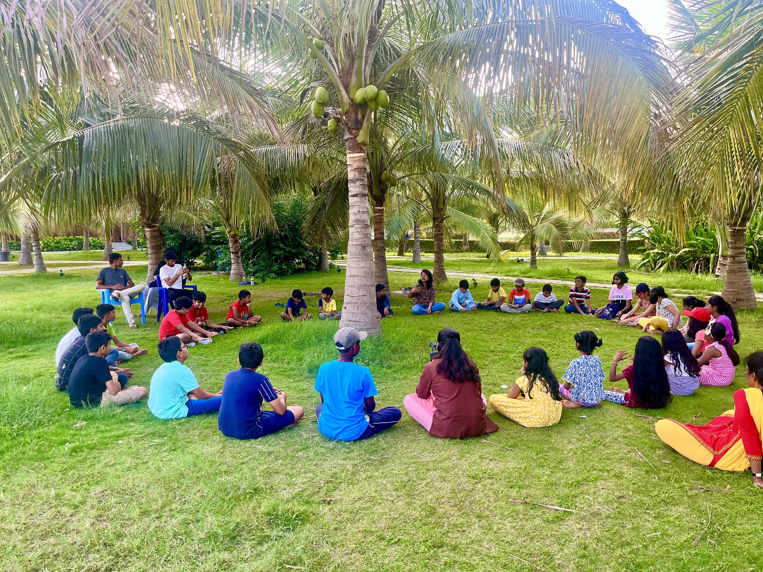 Children and adults sitting in a circle on the grass under palm trees in a park.