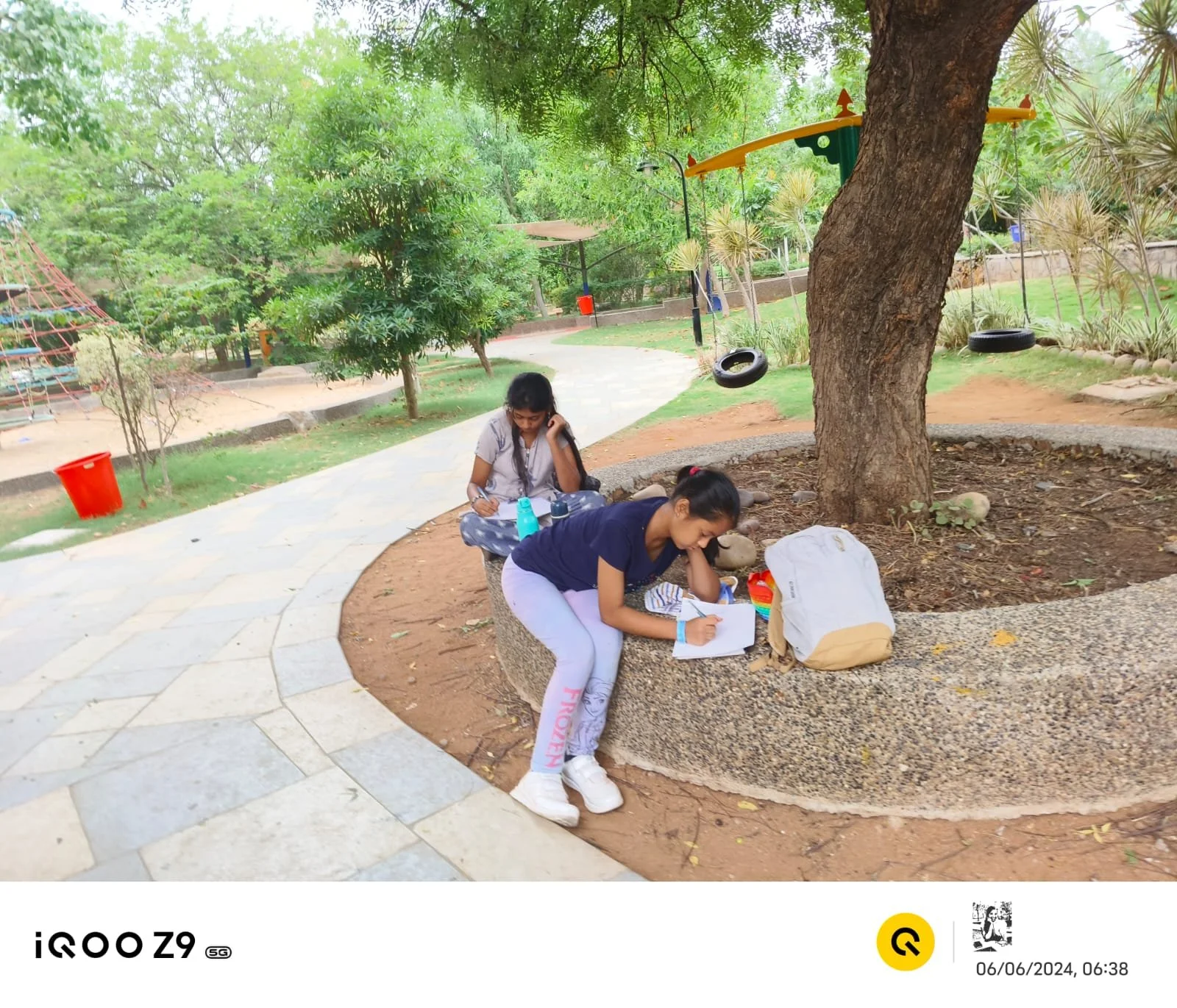 Two young women are sitting on a stone ledge under a large tree in a park. One woman is writing in a notebook, and the other is looking at her phone. There are backpacks and water bottles nearby, with children’s play equipment and trees visible in th