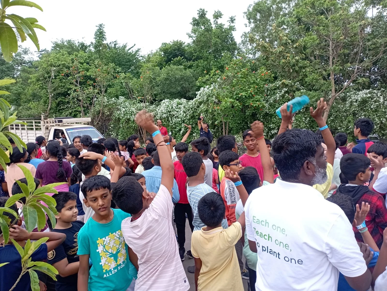 Children and adults gathered outdoors, some raising hands, with a vehicle and lush greenery in the background.