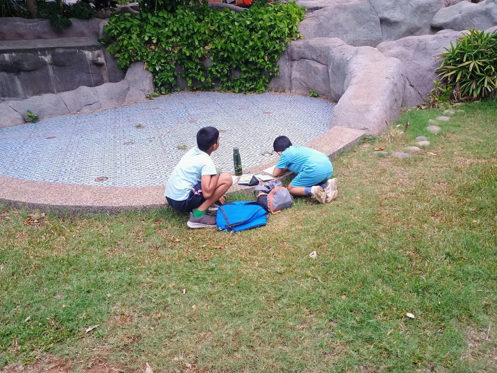 Two boys in casual clothes kneeling and sitting on the grass next to a small empty pond, surrounded by rocks and greenery, with backpacks and bottles nearby.