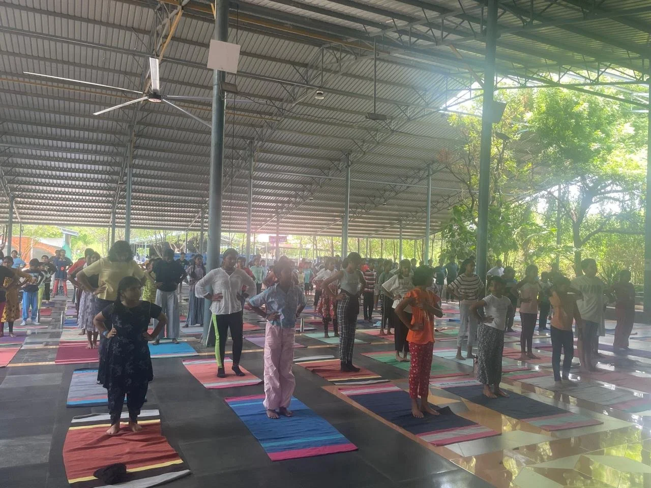 People participating in a group yoga class under a large open-air shelter with a metal roof, with trees and sunlight in the background.