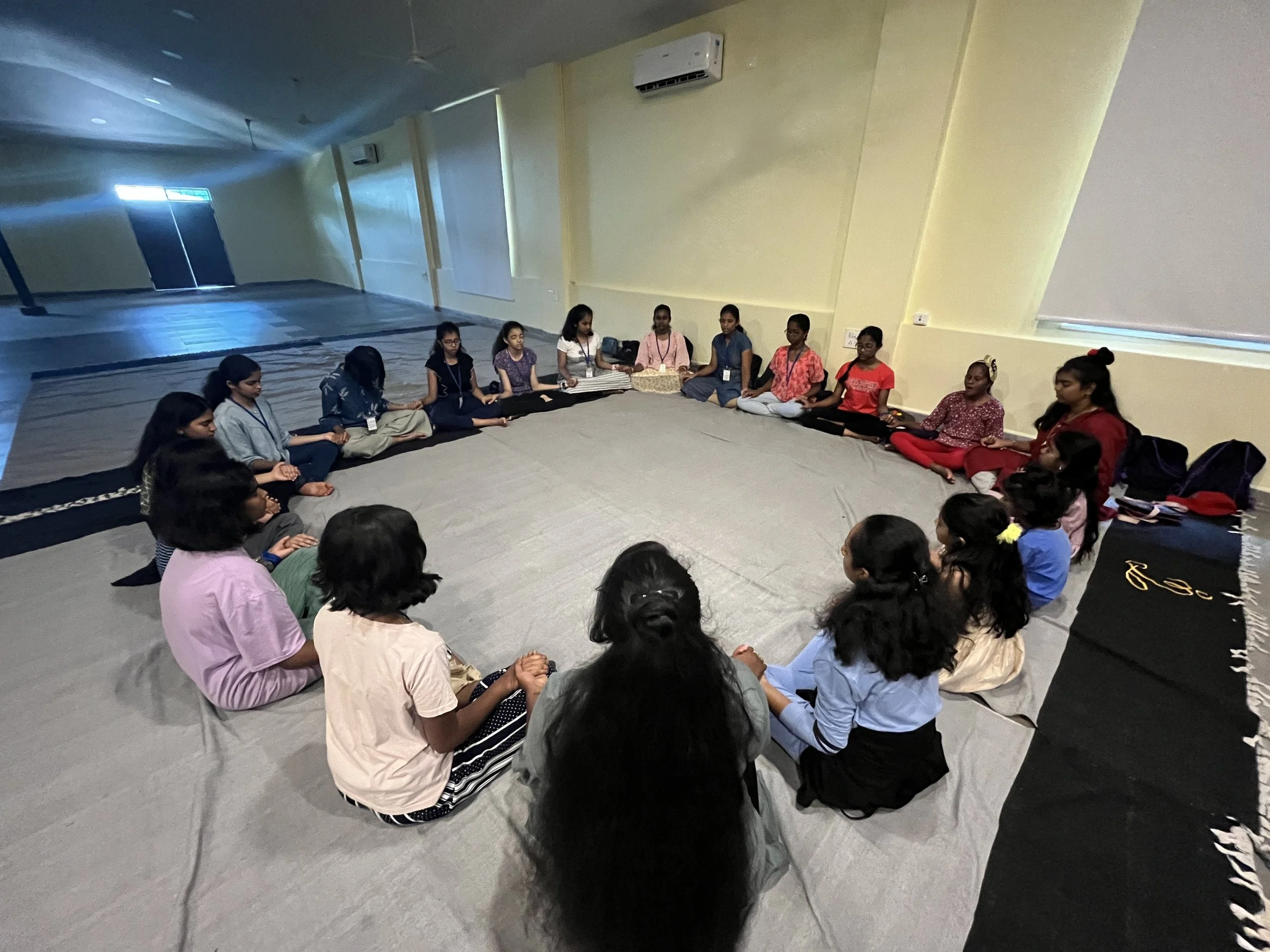 A group of young girls sitting cross-legged in a circle on a carpeted floor inside a room, engaged in a meditation or group activity.